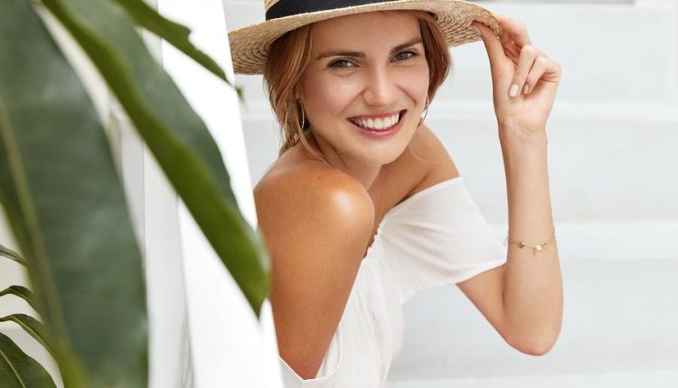 Sideways portrait of pleased happy woman wears summer straw hat, demonstrates bare shoulders, sits in hotel room, has appealing appearance and positive smile. People, leisure and joy concept